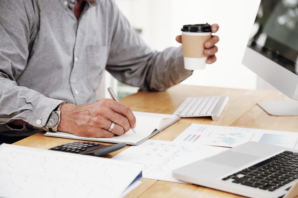 Close-up of businessman writing plans in his notepad at desk and holding cup of coffee in his hand