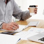 Close-up of businessman writing plans in his notepad at desk and holding cup of coffee in his hand