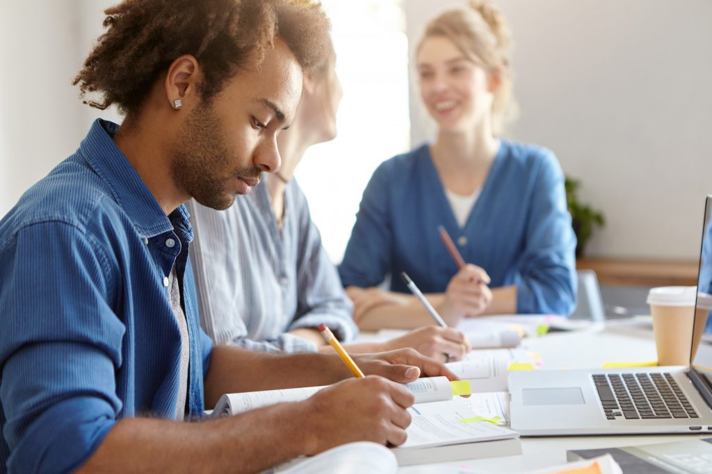 Stylish dark-skinned male in blue shirt, being busy with studying, sitting near his female groupmates, working laptop computer, writing diploma paper. Group of friendly students of different races